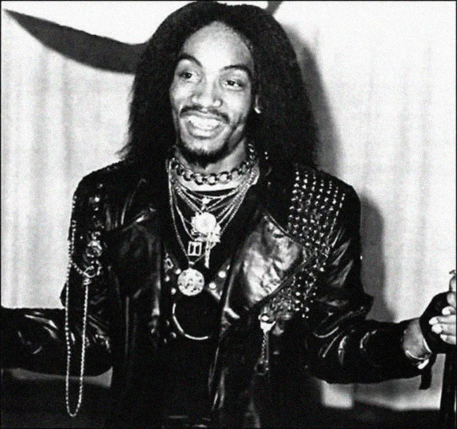 Black and white photo of a smiling person with long hair, wearing a studded leather jacket, layered necklaces, and jewelry, standing with arms outstretched against a plain background.