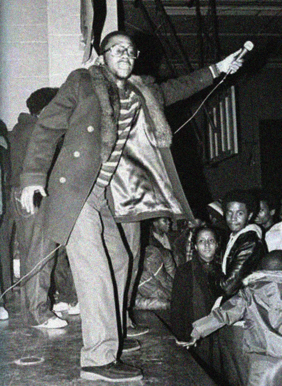 A man in glasses and a fur-collared coat stands on stage holding a microphone, engaging an audience below. Several people watch him from the crowd and stage, some smiling, in a lively indoor setting.
