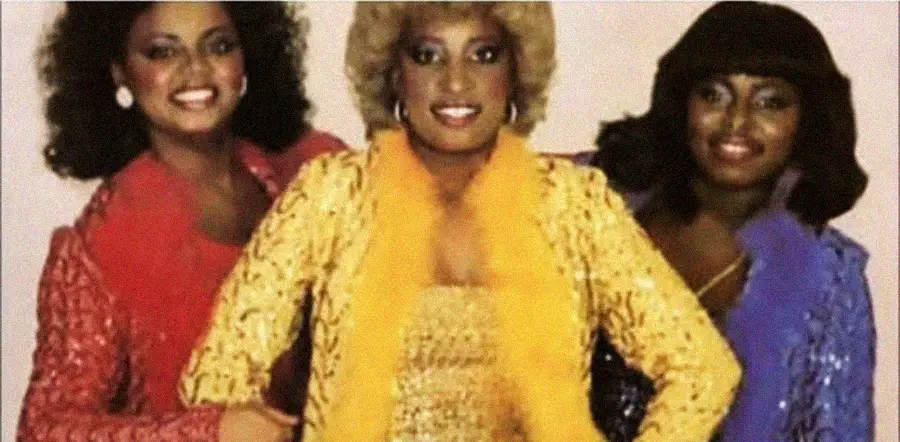 Three women pose together, smiling and dressed in sparkling outfits—red, gold, and purple—with matching feathered trims. They have styled hair and glamorous makeup, standing close against a plain background.