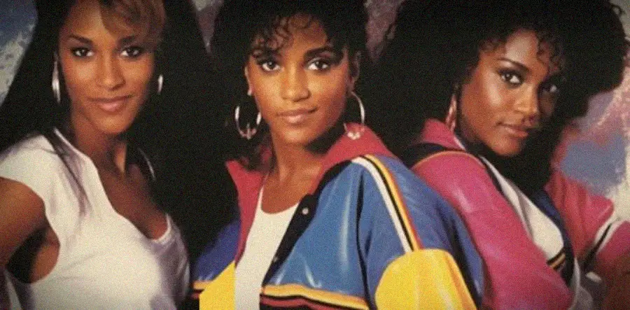 Three women pose closely together, all with curly hair and hoop earrings. The woman in the center wears a colorful jacket, while the others wear white and pink tops. The background is a soft, abstract pattern.