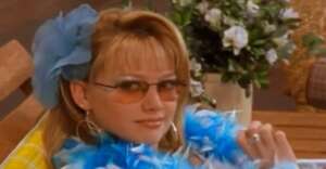 A person with blonde hair, wearing tinted sunglasses, a blue feather boa, large hoop earrings, and a blue flower hair accessory, sits on a wooden bench with flowers in the background.