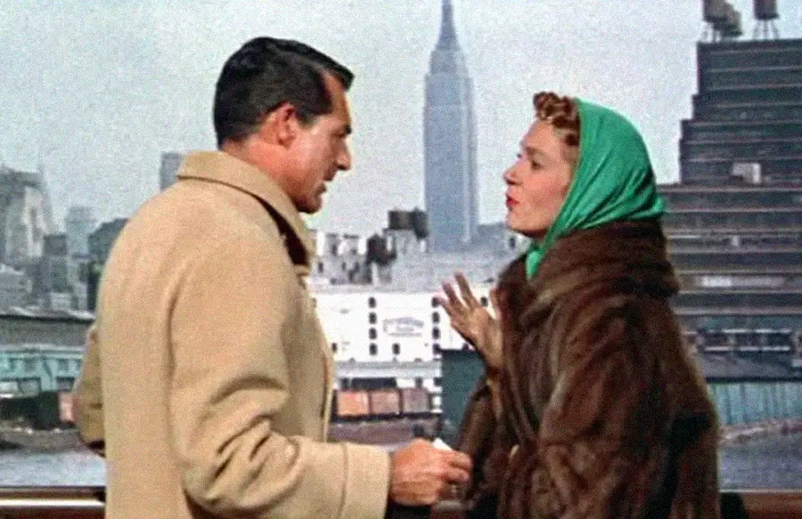 A man in a beige coat and a woman in a fur coat with a green scarf talk on a city rooftop with the Empire State Building visible in the background.