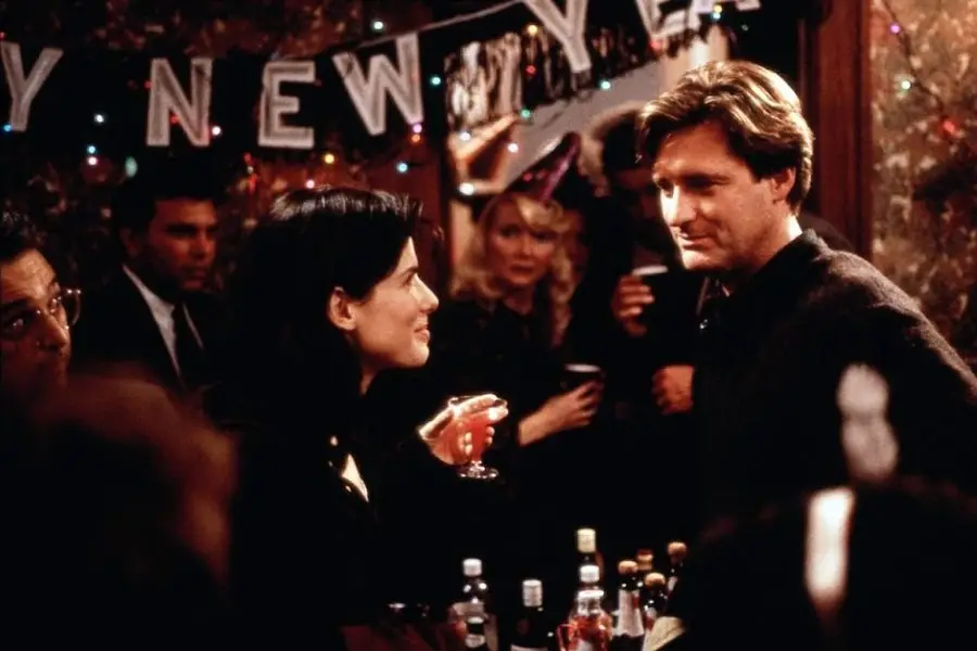 A woman holding a cocktail and a man stand talking at a decorated bar during a New Year’s Eve party, with a "Happy New Year" banner and people celebrating in the background.