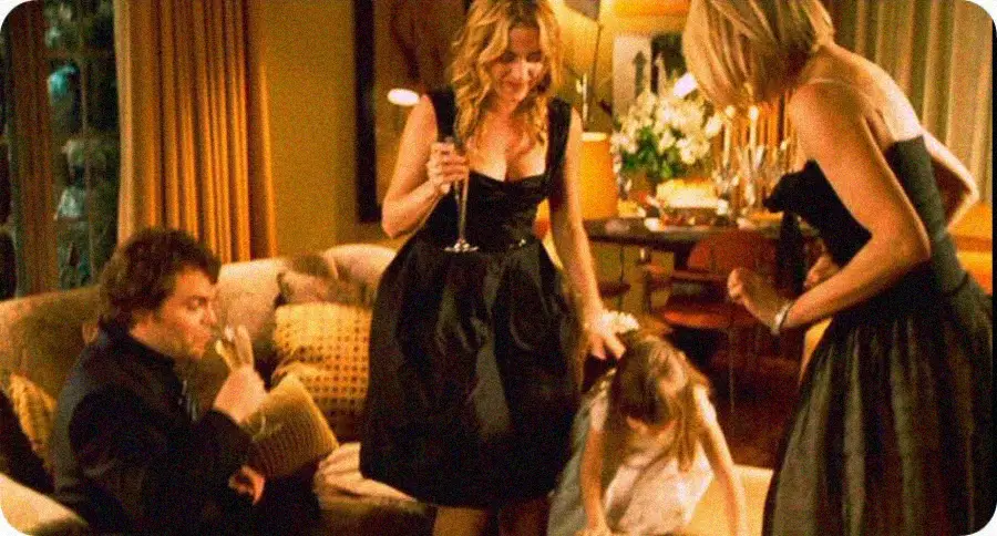 Three adults dressed formally and a child are in a warmly lit living room. One man sits on a couch drinking from a glass, while two women stand, one holding a drink, as a young girl crouches on the floor in front of them.