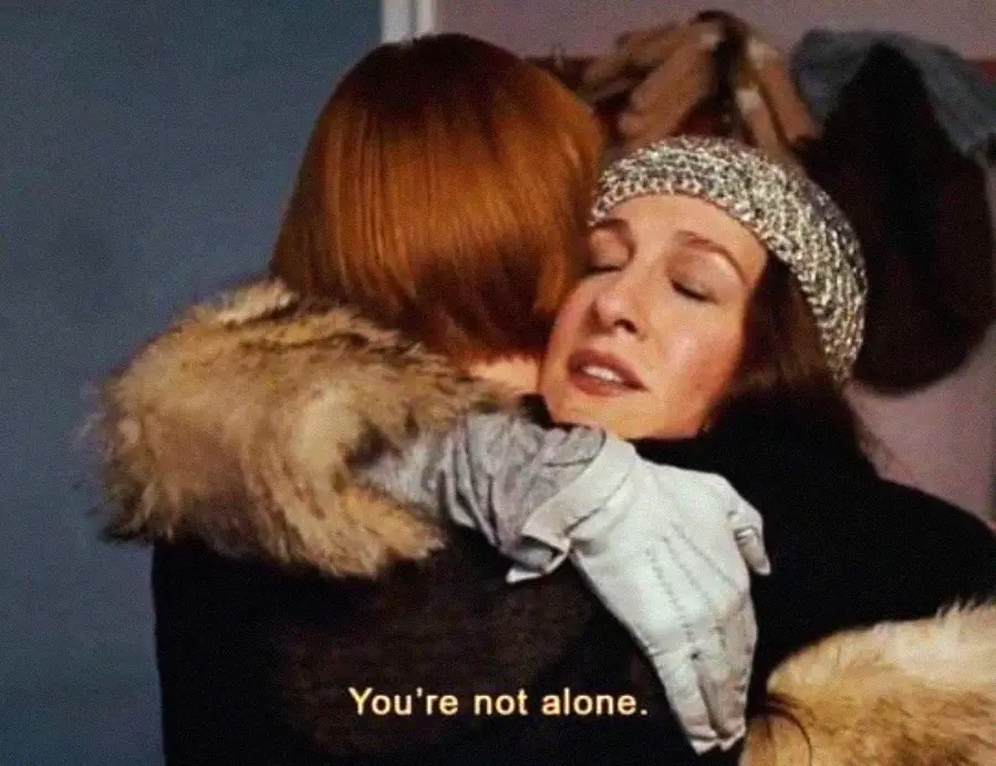 Two women are hugging each other tightly, one wearing a sparkly headband and a fur coat. The caption on the image reads, "You're not alone.