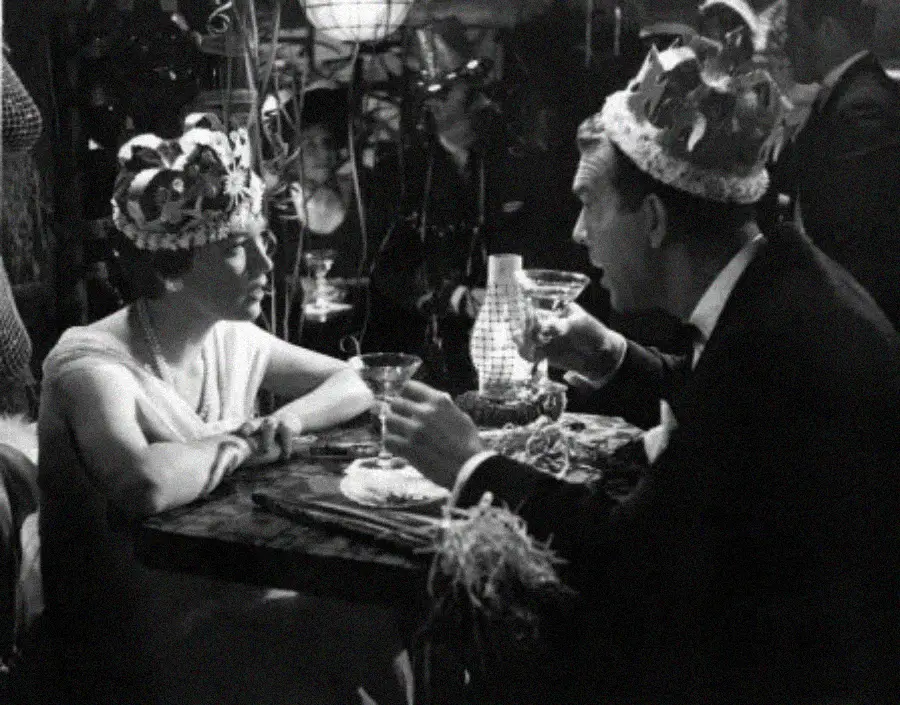 A woman and man wearing festive crowns sit at a small table, holding drinks and talking at a lively party, with decorations and other people visible in the background.
