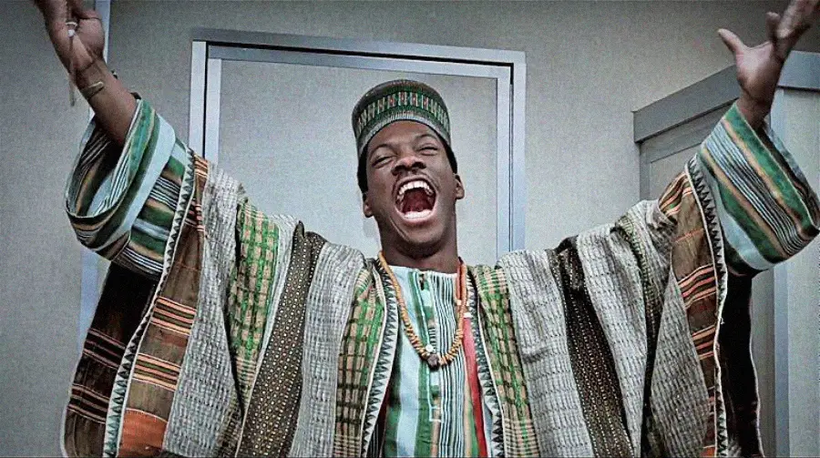 A man wearing a colorful traditional African robe and hat stands indoors with arms raised, smiling widely and appearing joyful.