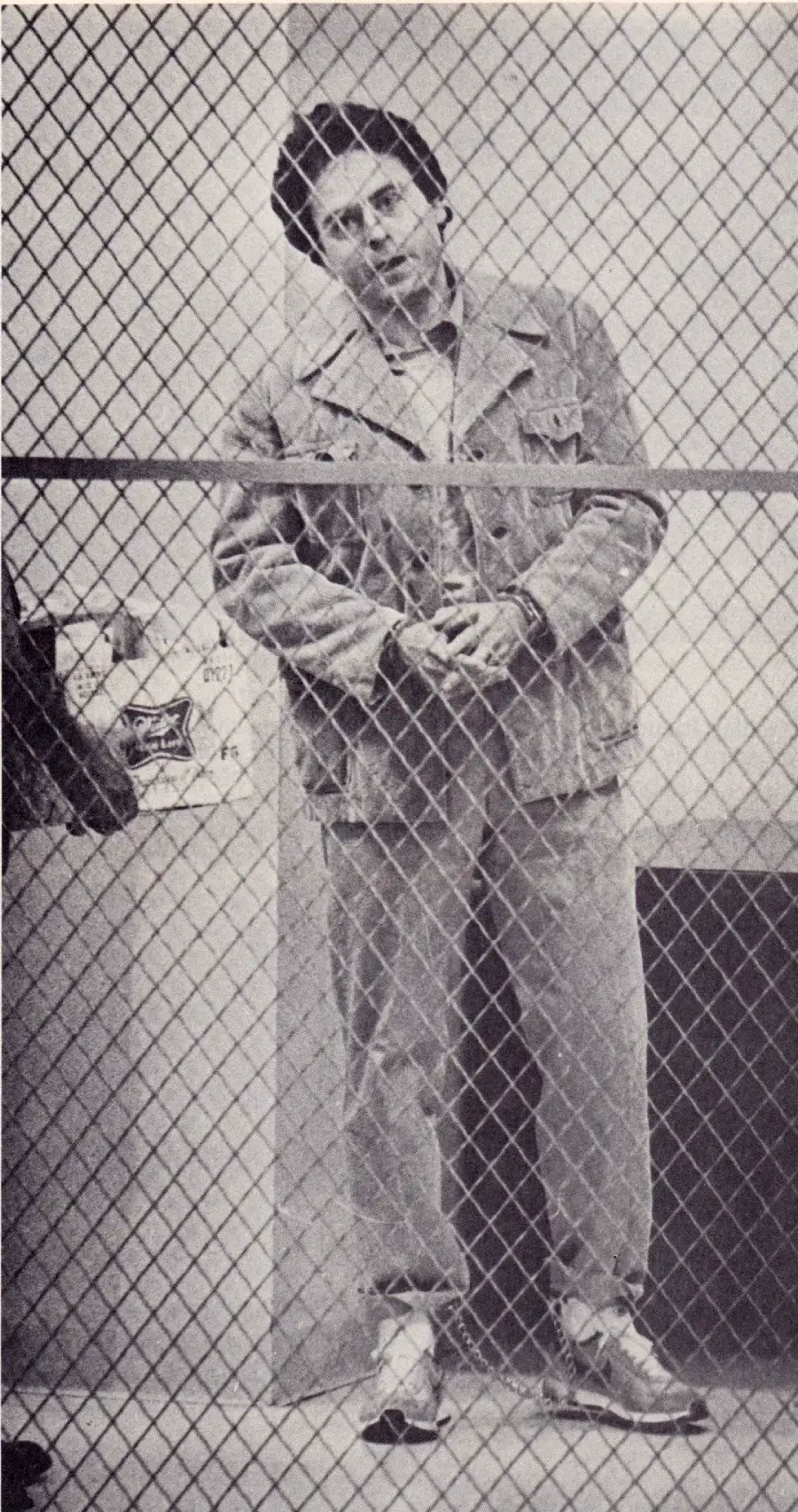 A person stands behind a chain-link fence, wearing glasses, a jacket, and casual pants. Their hands appear to be restrained in front, and they look slightly to the side in a confined space.
