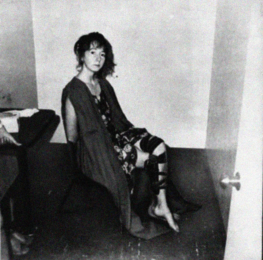 A woman in a patterned dress and robe sits barefoot on the floor against a wall, her hands behind her back and legs restrained with straps in a sparse, dimly lit room.