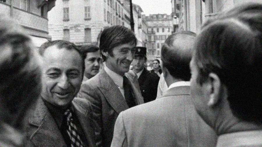 A group of men in suits stand closely together on a city street, smiling and talking. The photo is black and white, capturing a candid moment in an urban setting with buildings in the background.