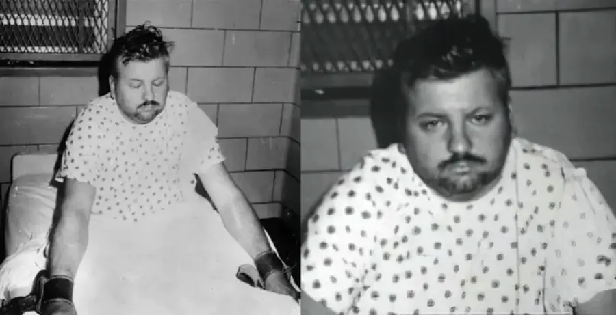 A man with dark hair and a mustache, wearing a hospital gown, sits on a bed in a cell-like room. His wrists are restrained, and he appears tired or somber. The photo is in black and white.