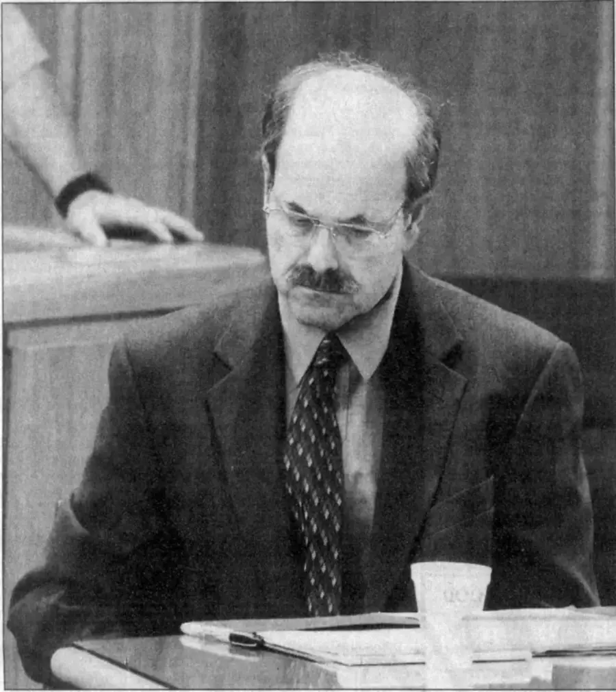 A middle-aged man with glasses, a mustache, and balding hair sits in a courtroom at a table, looking down with a serious expression. He wears a suit and tie; a paper and cup are on the table.