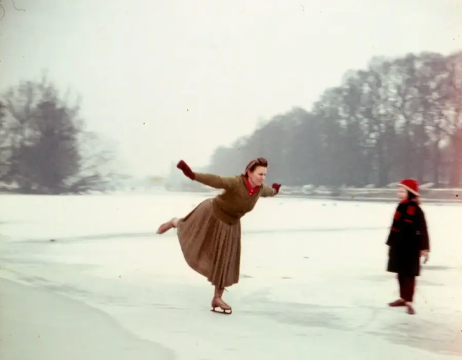 A woman ice skating outdoors with one leg raised behind her, wearing a brown skirt, red scarf, and red mittens. A child in a dark coat and red hat stands nearby. Snow covers the ground and trees are in the background.