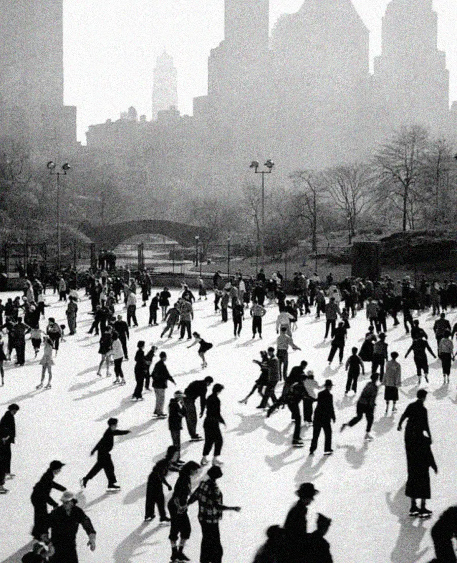 Black-and-white photo of a crowded ice skating rink in a city park, with people skating and walking. Leafless trees and tall buildings fill the background, creating a wintry urban scene.
