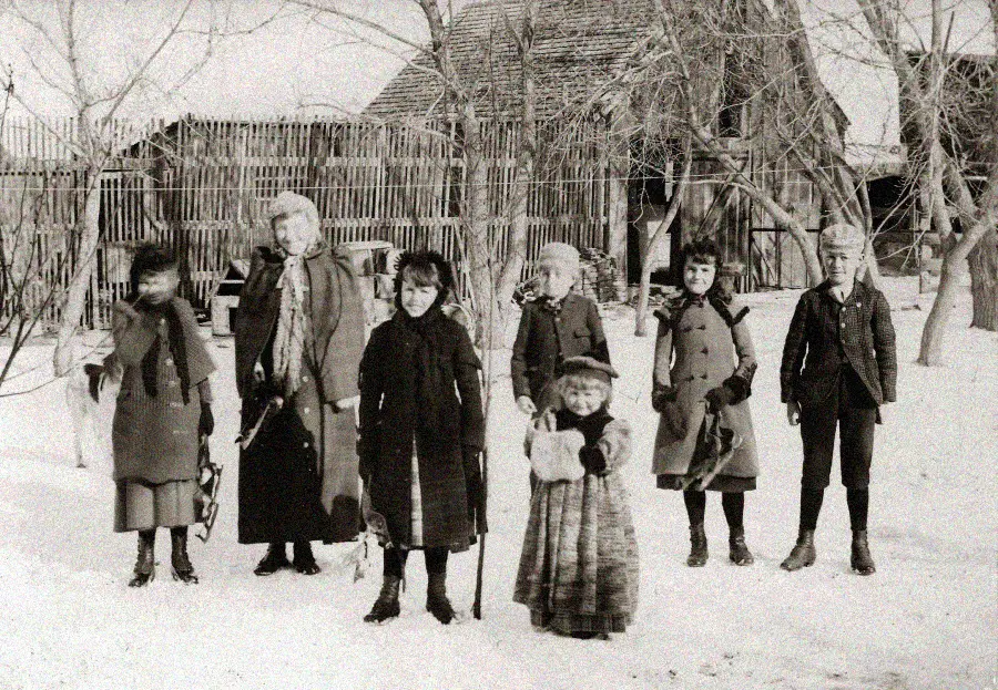 A vintage black-and-white photo of six children and one woman standing outside in the snow, dressed in winter clothing, with wooden fences and buildings in the background.