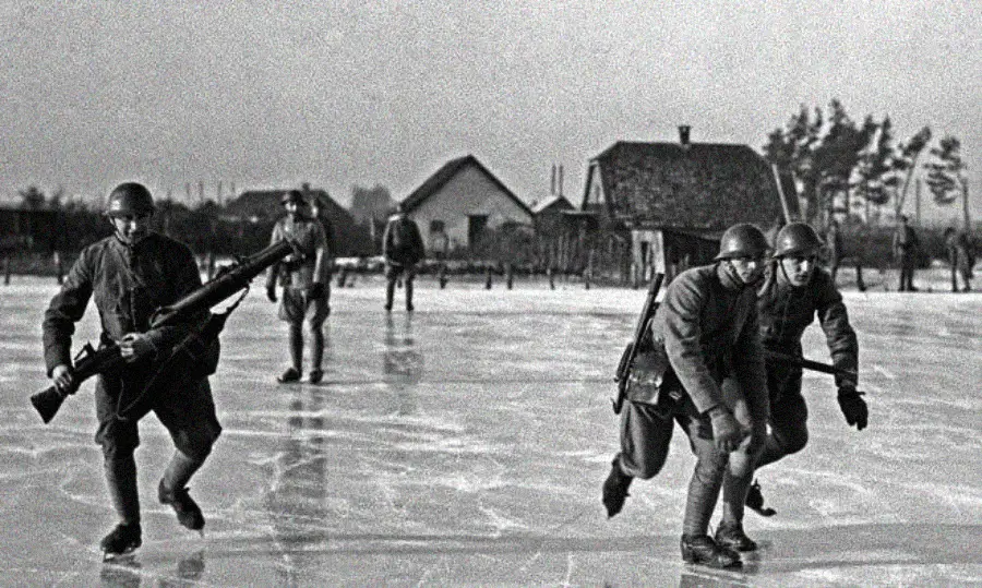 Soldiers in uniform and helmets cross a frozen, icy surface, some carrying rifles. Houses and trees are visible in the background under an overcast sky.