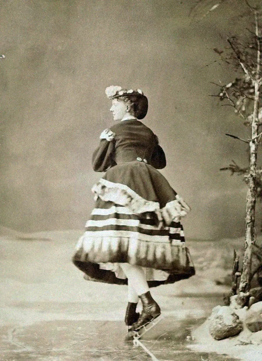 A vintage black-and-white photo of a woman ice skating. She wears a long, layered dress with a bustle and a floral hat, looking away from the camera. Leafless trees and snow are seen in the wintry background.
