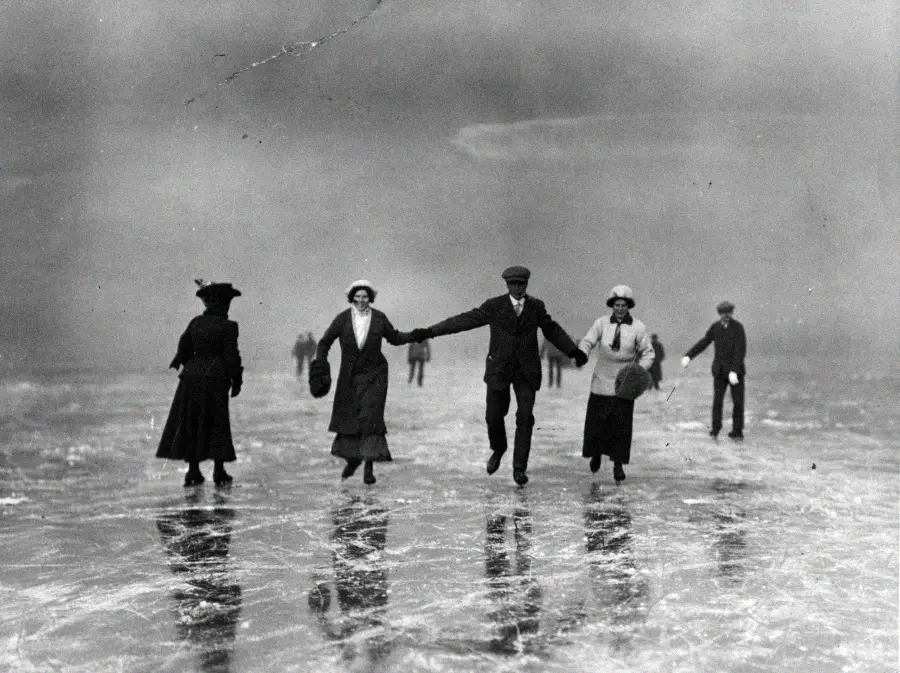 A black-and-white photo of people ice skating on a frozen lake. Three women and a man in early 20th-century clothing skate together, holding hands, while two others skate behind them. Their reflections are visible on the ice.
