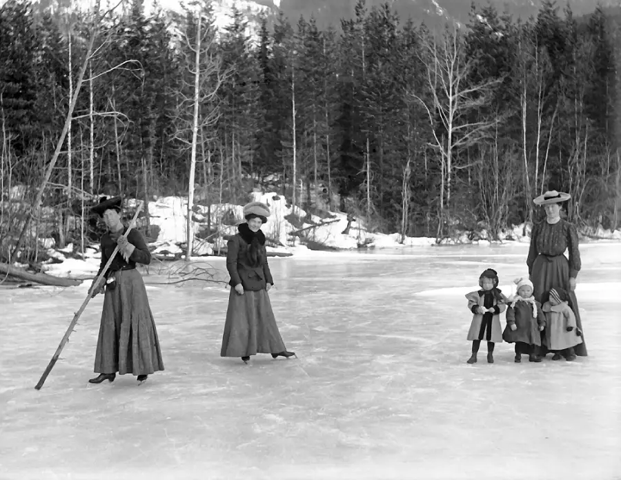 Three women in long dresses and hats stand on a frozen lake, with two guiding four small children on the ice. Leafless trees and a forested area are in the background, suggesting wintertime.