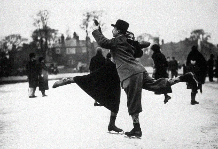 A vintage black and white photo of a man and woman figure skating on an outdoor ice rink, striking a graceful pose with one leg extended, while other people skate and watch in the background.