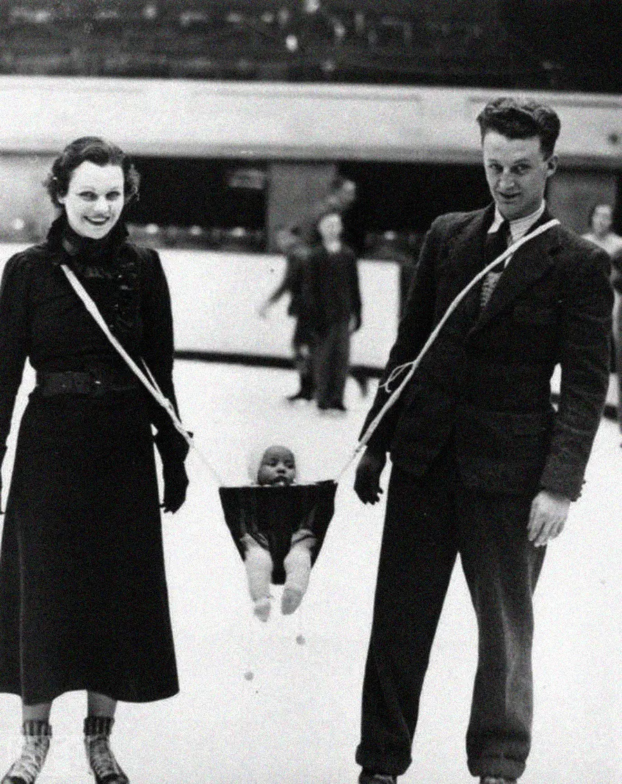 A smiling woman and man ice skate while holding a sling between them that supports a baby, also on the ice. People are visible skating in the background. The image appears to be from the early to mid-20th century.