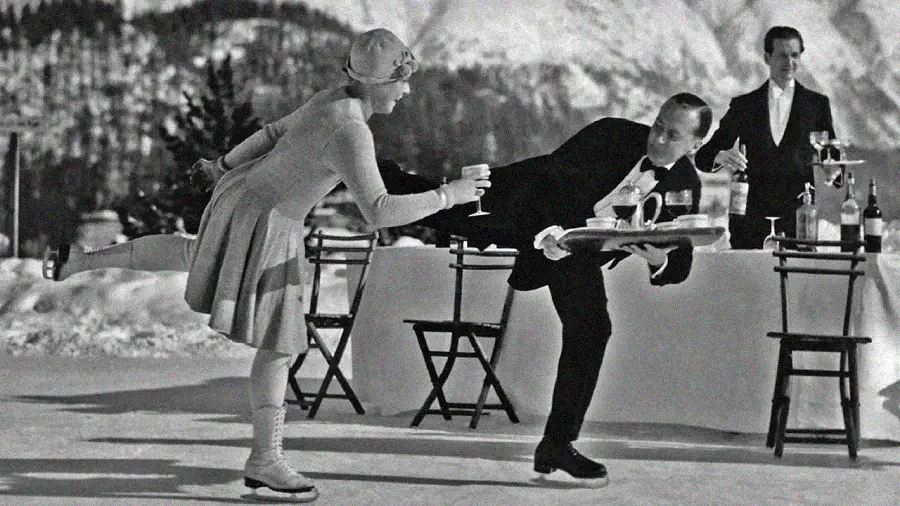 A woman and a waiter, both ice skating outdoors, balance skillfully as she takes a drink from his tray. Another waiter and empty chairs are visible behind them, with snowy mountains in the background.