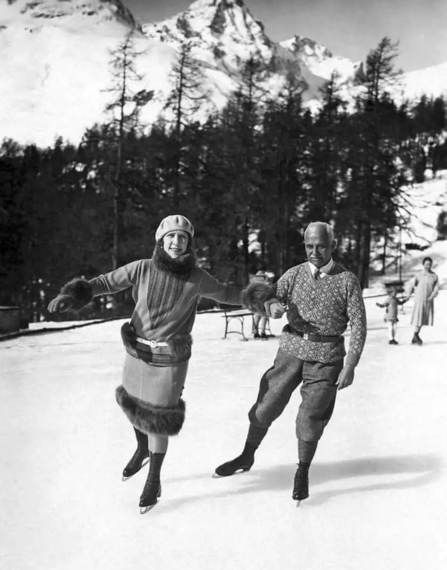 A smiling woman and man ice skate outdoors on a snowy day, dressed in vintage winter clothing, with mountains and trees in the background and other skaters visible.