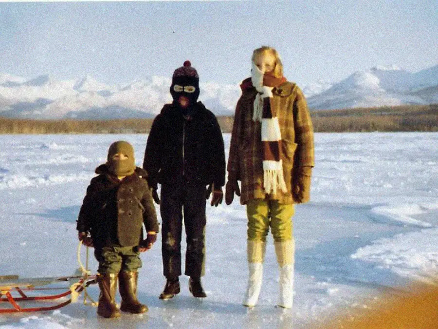 Three people, warmly dressed in winter clothing and masks, stand on a frozen lake with snowy mountains in the background. A small sled is beside the child on the left.