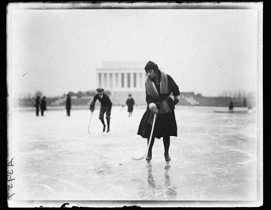 A woman in vintage winter clothing plays hockey on an ice rink near the Lincoln Memorial, while others skate in the background on a foggy day.