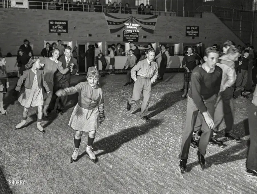 A group of boys and girls ice skating indoors, some holding hands and others skating solo. People watch from the stands above, and signs on the wall display skating rules. Everyone is warmly dressed.
