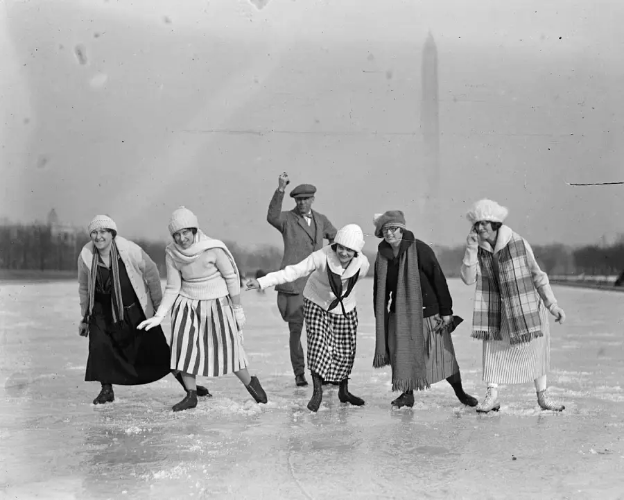Six people, five women and one man, wearing winter clothes and hats, ice skate on an outdoor rink with the Washington Monument visible in the foggy background. They are smiling and posing playfully.