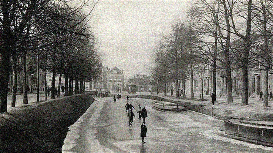 Black and white photo of people ice skating on a frozen canal lined with leafless trees and buildings on both sides, capturing a winter scene in a historic European city.