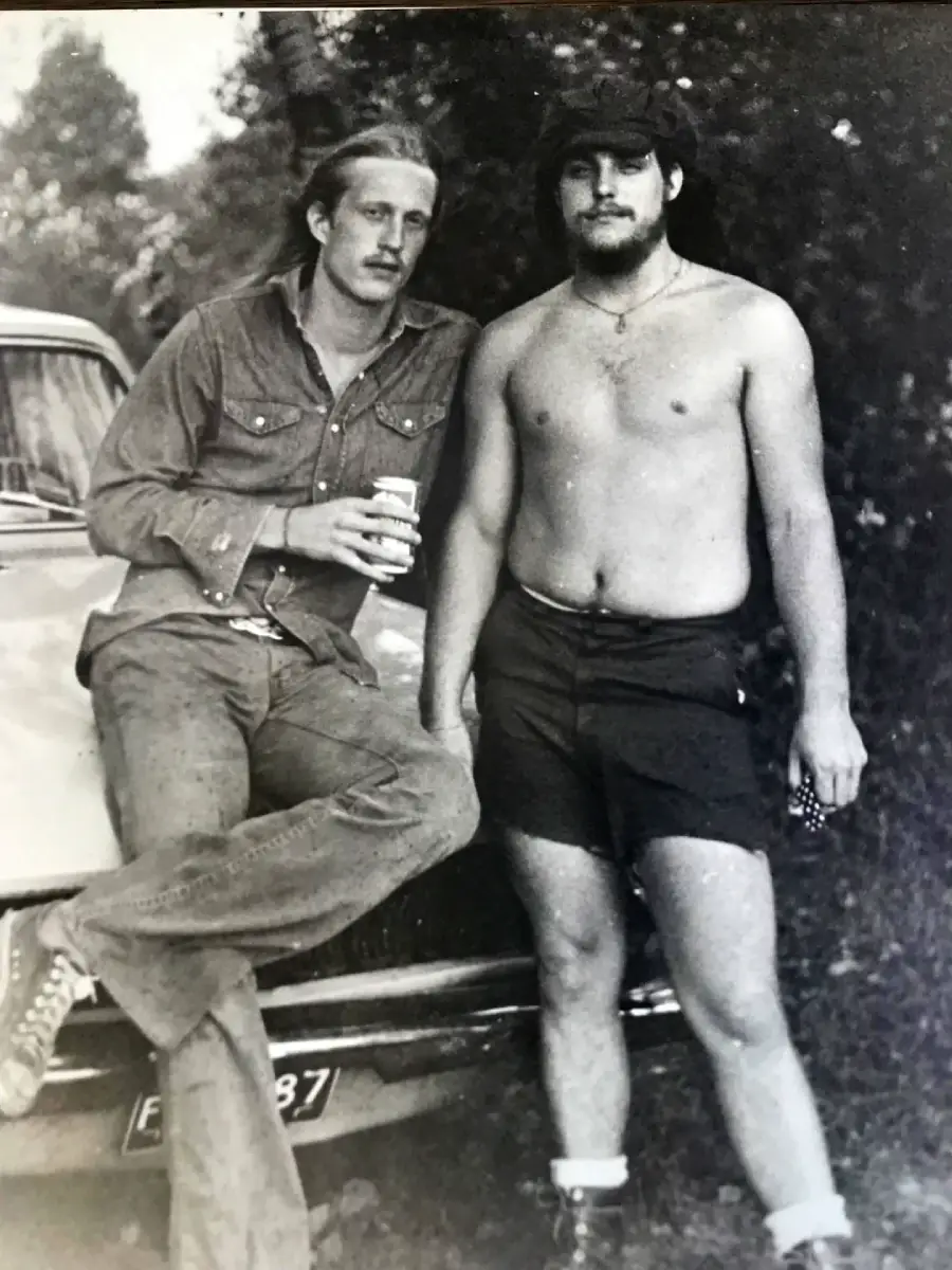 Two men pose beside a car; one is leaning against the hood holding a drink, wearing jeans and a buttoned shirt, while the other stands shirtless in shorts, both looking at the camera in a black and white outdoor setting.