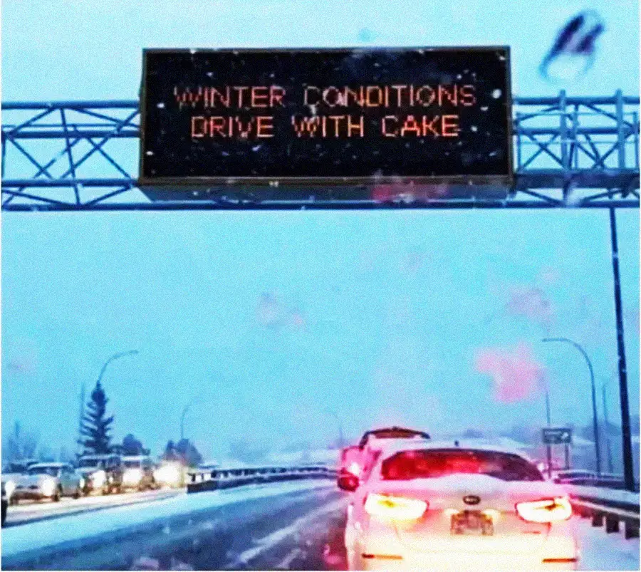 Cars drive on a snowy road under an electronic highway sign that mistakenly reads, "WINTER CONDITIONS DRIVE WITH CAKE." The sky is overcast and snow covers the ground and vehicles.