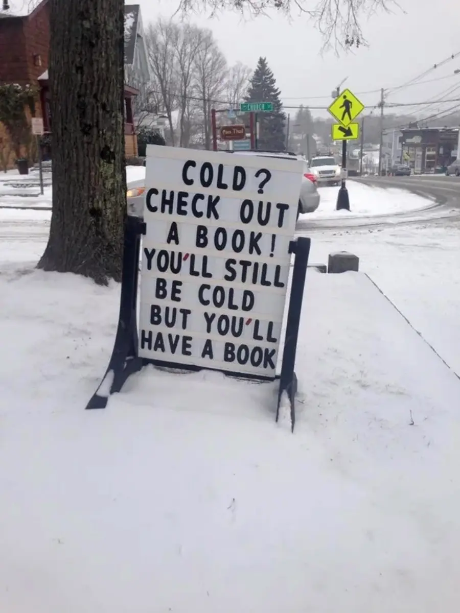 A sidewalk sign on snowy ground reads: “Cold? Check out a book! You’ll still be cold but you’ll have a book.” Cars and buildings are visible in the background on a wintry street.