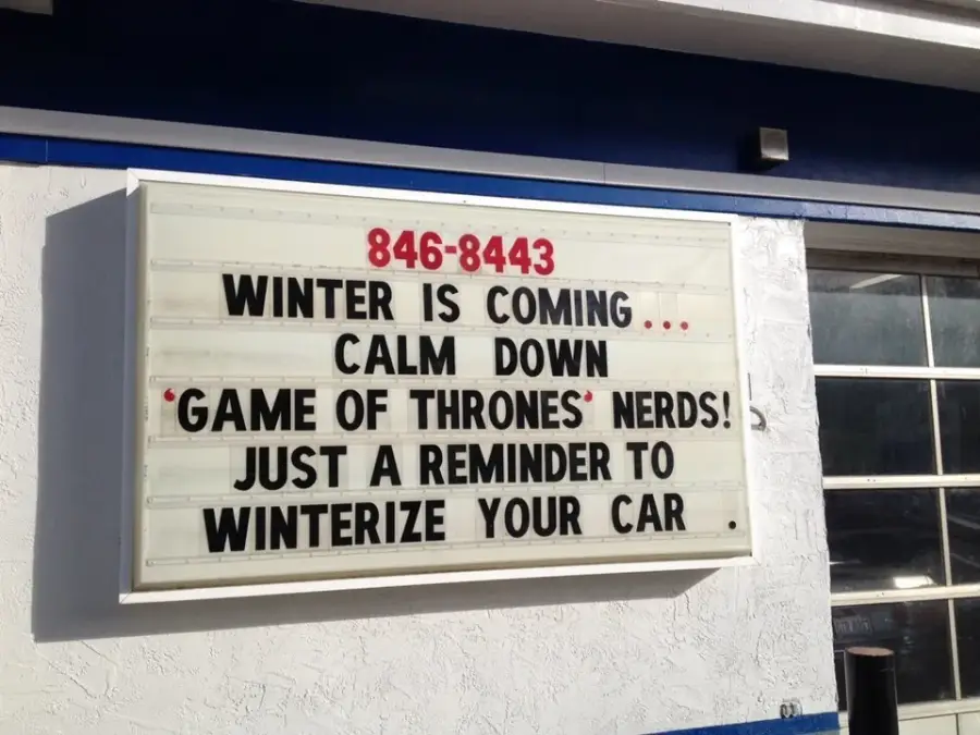 A sign reads: "846-8443 WINTER IS COMING... CALM DOWN 'GAME OF THRONES' NERDS! JUST A REMINDER TO WINTERIZE YOUR CAR." The sign is attached to a white and blue building.