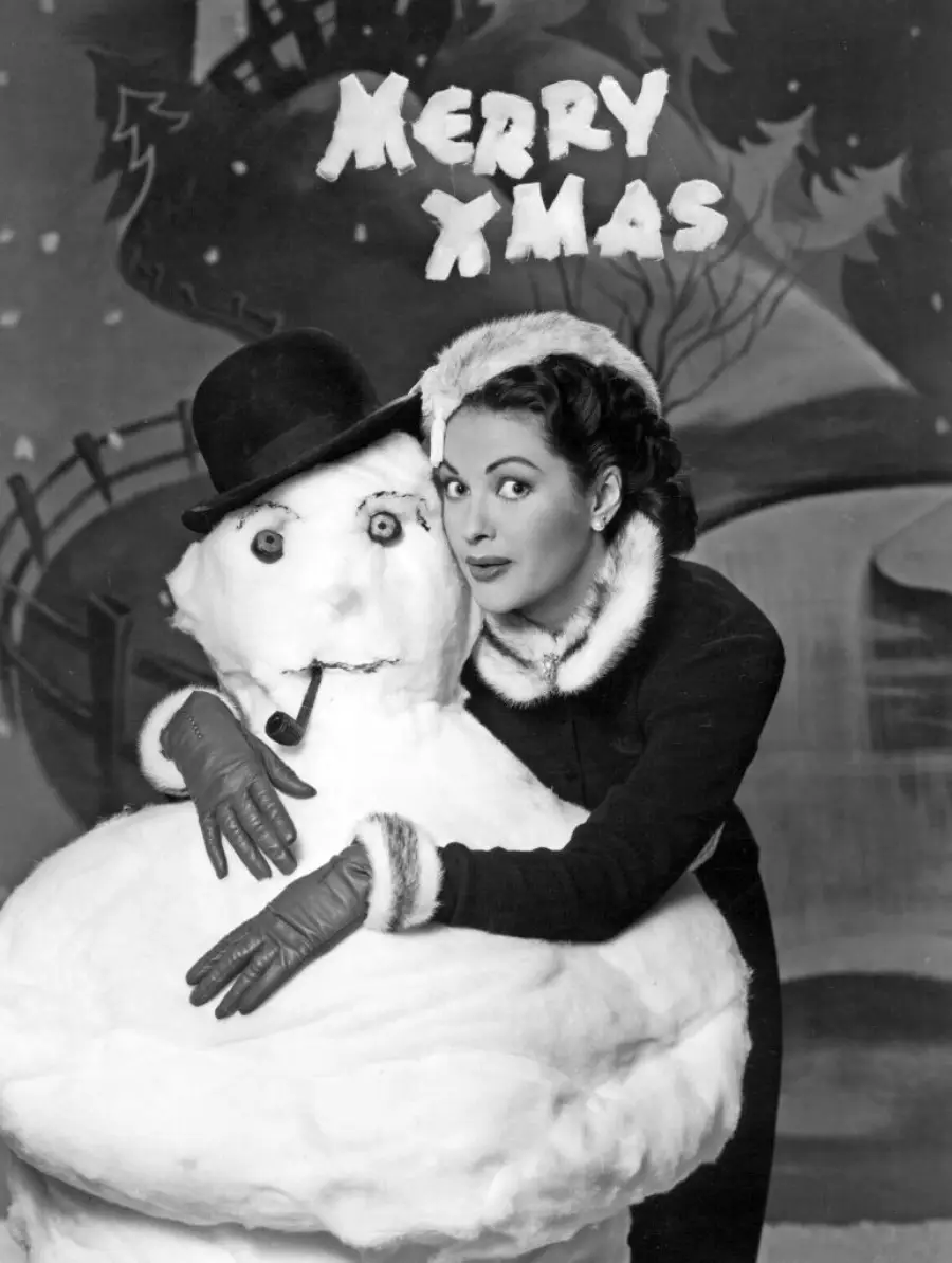 A woman in winter clothing poses with a snowman wearing a hat and pipe. Behind them, "MERRY XMAS" is written in festive letters amidst a snowy, winter scene.