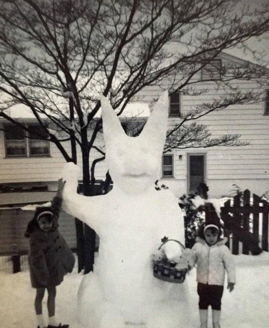 Two children in winter coats stand beside a large snow sculpture resembling a rabbit with tall ears, holding a basket. Snow covers the ground and the house behind them. The scene is black and white.