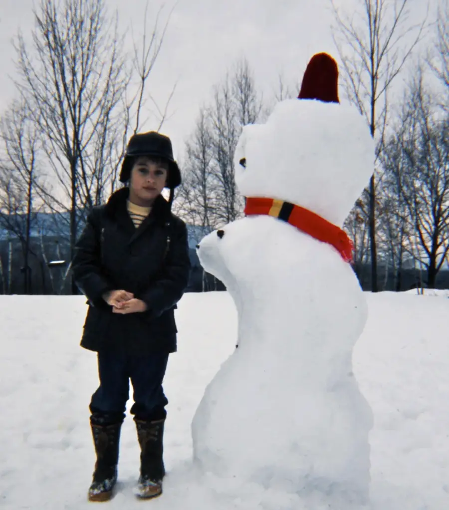 A young child in winter clothes and boots stands next to a tall snowman wearing a red scarf and red hat in a snowy park with leafless trees in the background.