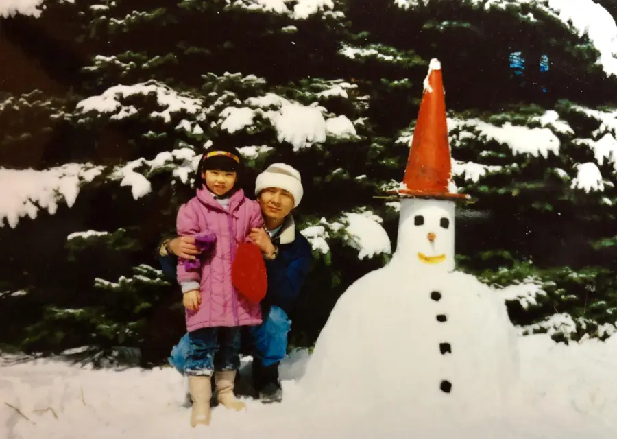 A man and a young girl in winter clothes stand next to a snowman with a red cone hat, orange nose, stick smile, and button eyes, in front of snow-covered evergreen trees.