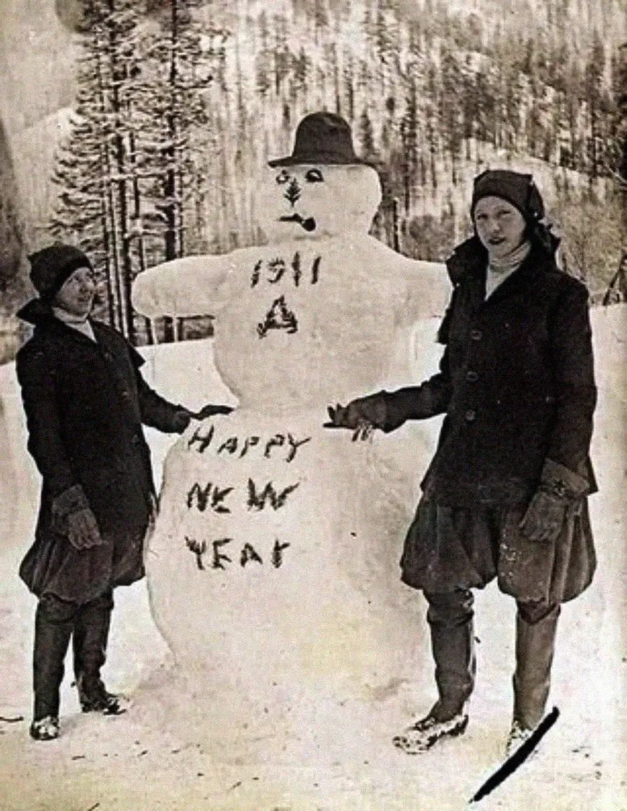 Two women in winter clothing stand next to a large snowman wearing a hat. The snowman has "1911 A Happy New Year" written on it. Snow-covered trees and hills are visible in the background.