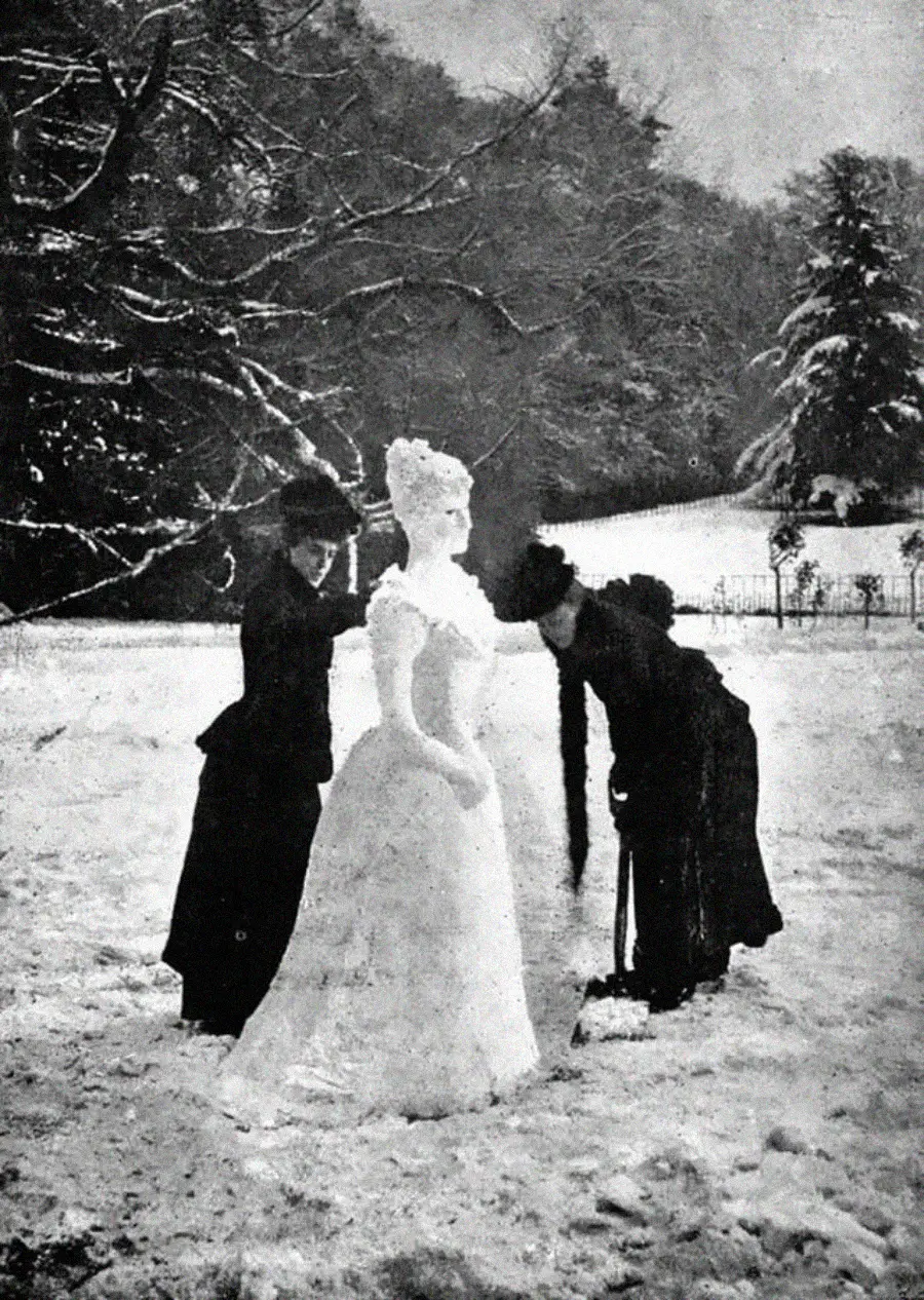 Two women in long dark dresses build an elaborate snowwoman with a tall hairstyle in a snowy park or garden, surrounded by trees and a fence in the background. The scene has an old-fashioned, vintage look.