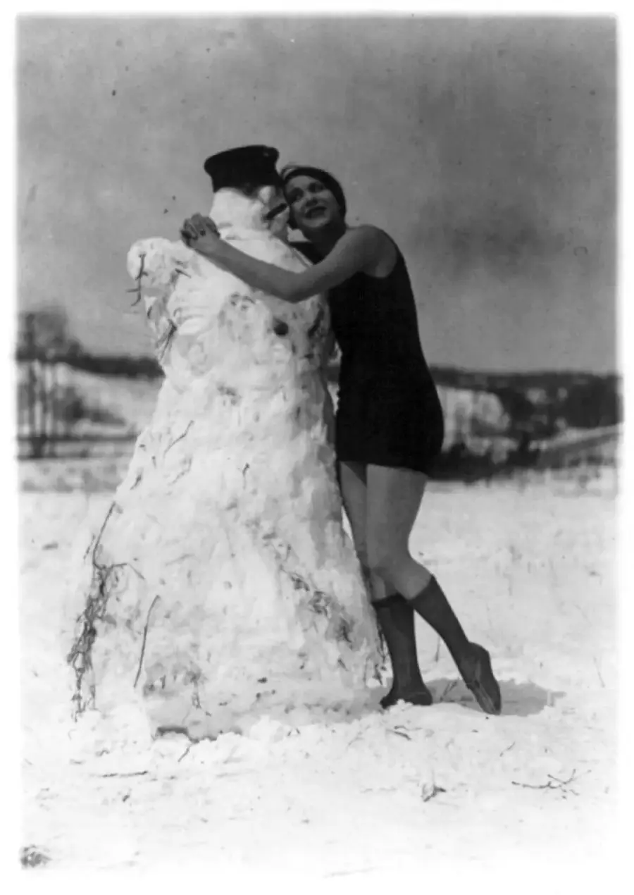 A woman in a short dress and tall socks hugs a large snowman wearing a dark hat in a snowy outdoor setting, with trees and hills visible in the background.