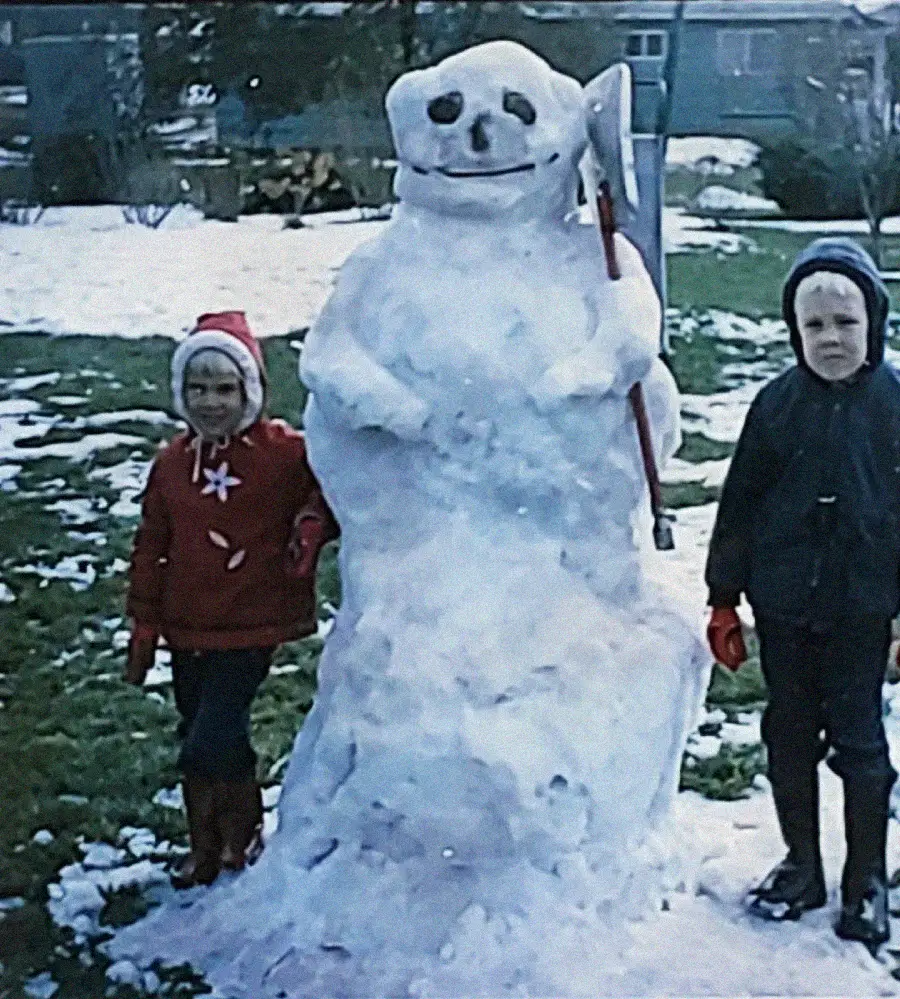 Two children stand outside in winter clothes next to a large snowman with a stick arm, button eyes, and a scarf, on a snowy lawn with houses and trees in the background.