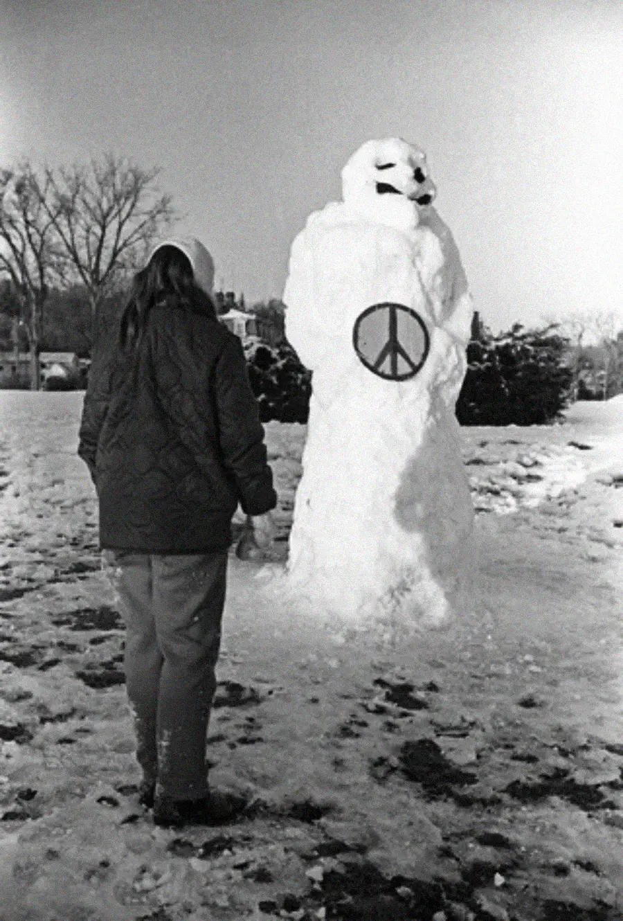 A person in winter clothing stands in front of a large snowman with a peace symbol drawn on its chest in a snowy outdoor setting.