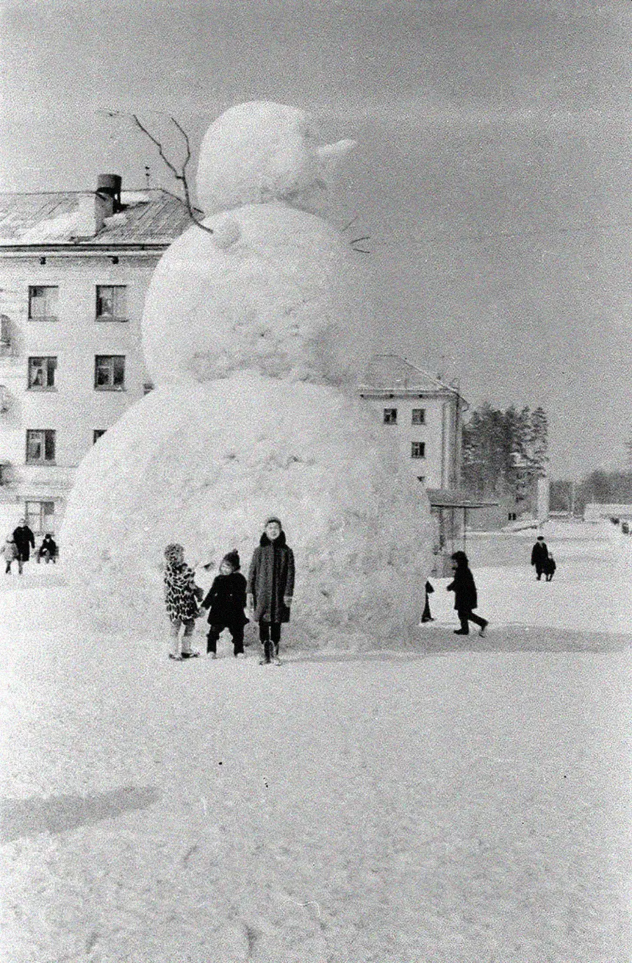 A group of people stand in front of a giant snowman with three large snowballs and stick arms, built in a snowy courtyard near multi-story buildings, with more people scattered in the background.