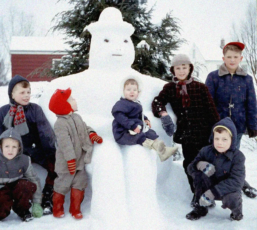 Six children in winter clothes pose around a large snowman with a hat, sitting in front of snow-covered trees and houses on a snowy day. One child sits on the snowman's lap.
