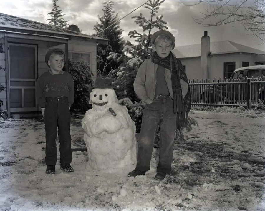 Two boys stand outside in the snow next to a snowman wearing a hat and scarf in front of a house. One boy is smiling and the other looks serious. Trees and a vintage car are visible in the background.