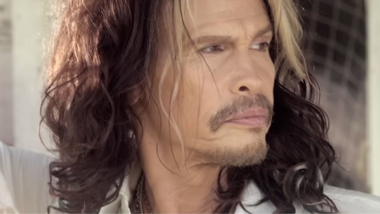 A person with long wavy brown hair and blonde highlights looks thoughtfully to the side, wearing a white shirt and necklace, with a soft light illuminating their face.