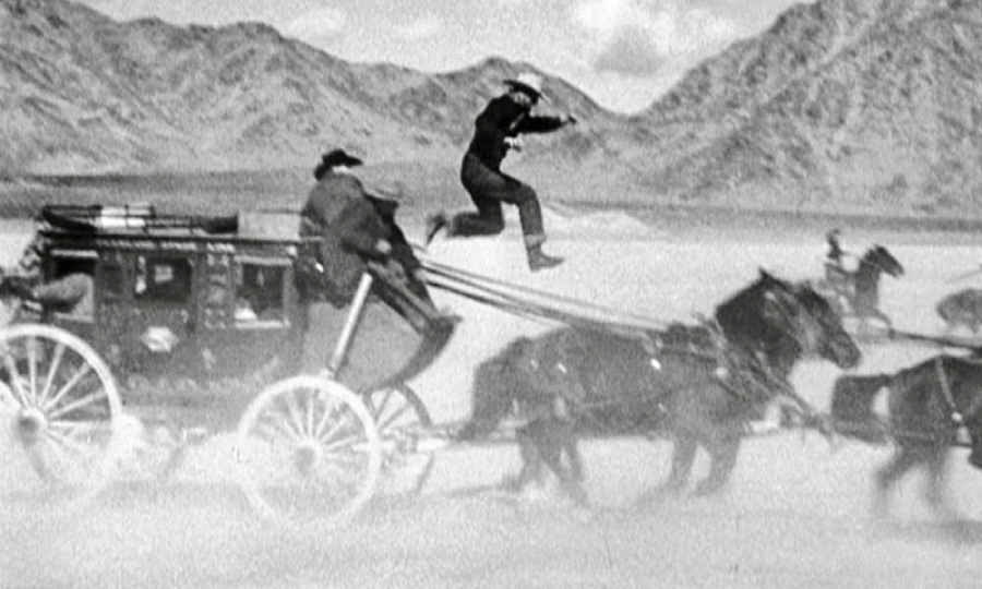 A person jumps from the top of a speeding stagecoach pulled by horses, while another person holds the reins; mountains and desert are in the background.